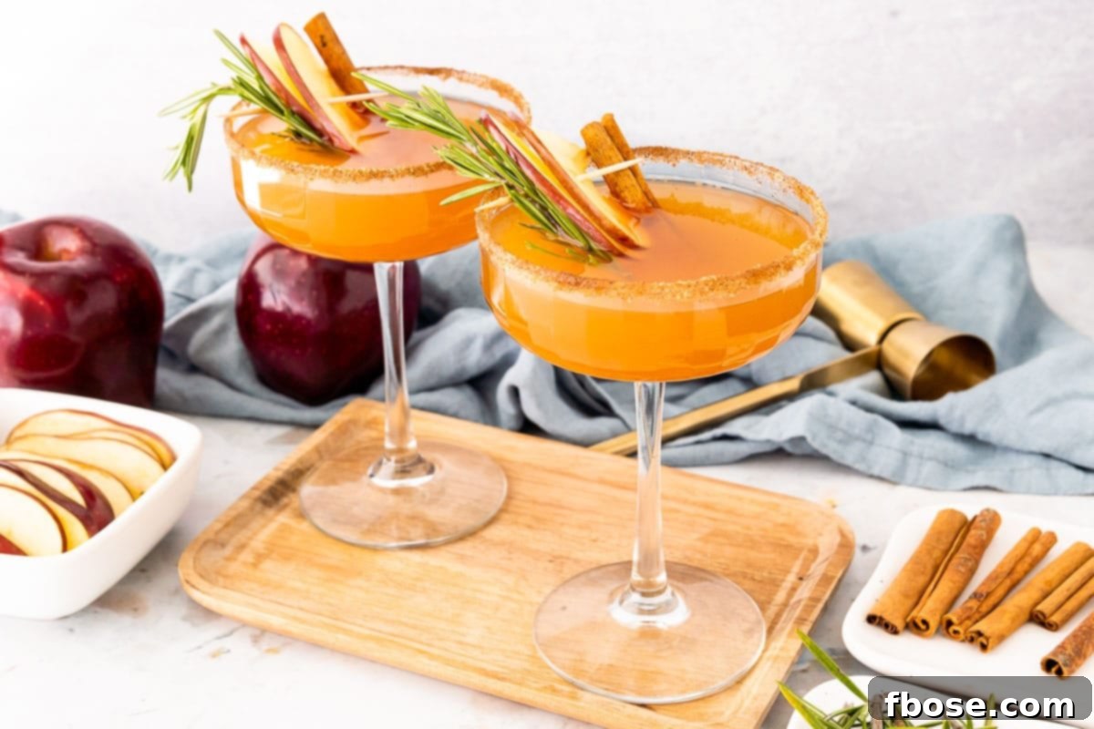 Fireside Apple Bourbon Elixir 5 Two Apple Cider Bourbon Cocktails in coupe glasses on a wooden board, garnished with apple slices and cinnamon sticks, ready to be served.
