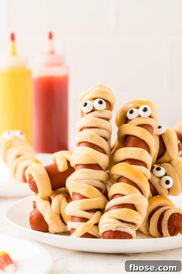 Mummy hot dogs on a white plate with mustard and ketchup in the background, showing their golden-brown perfection.