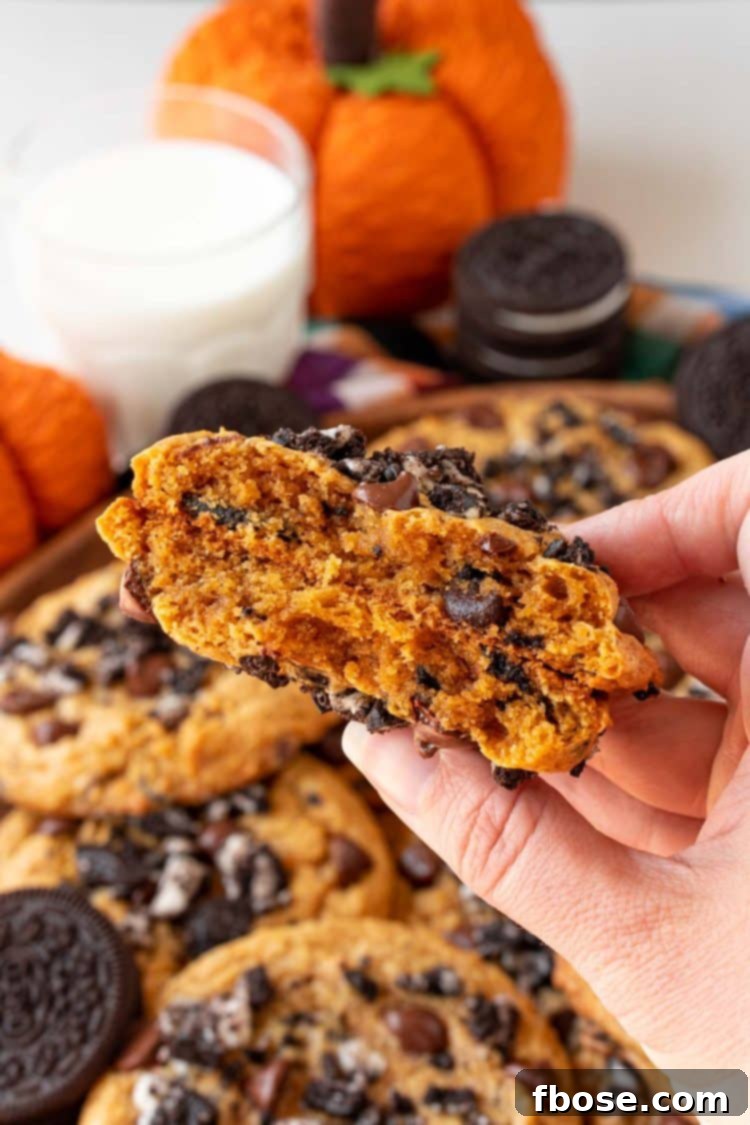 A woman's hand holding a pumpkin chocolate chip cookie with oreos in it broken in half so you can see the chewy center.