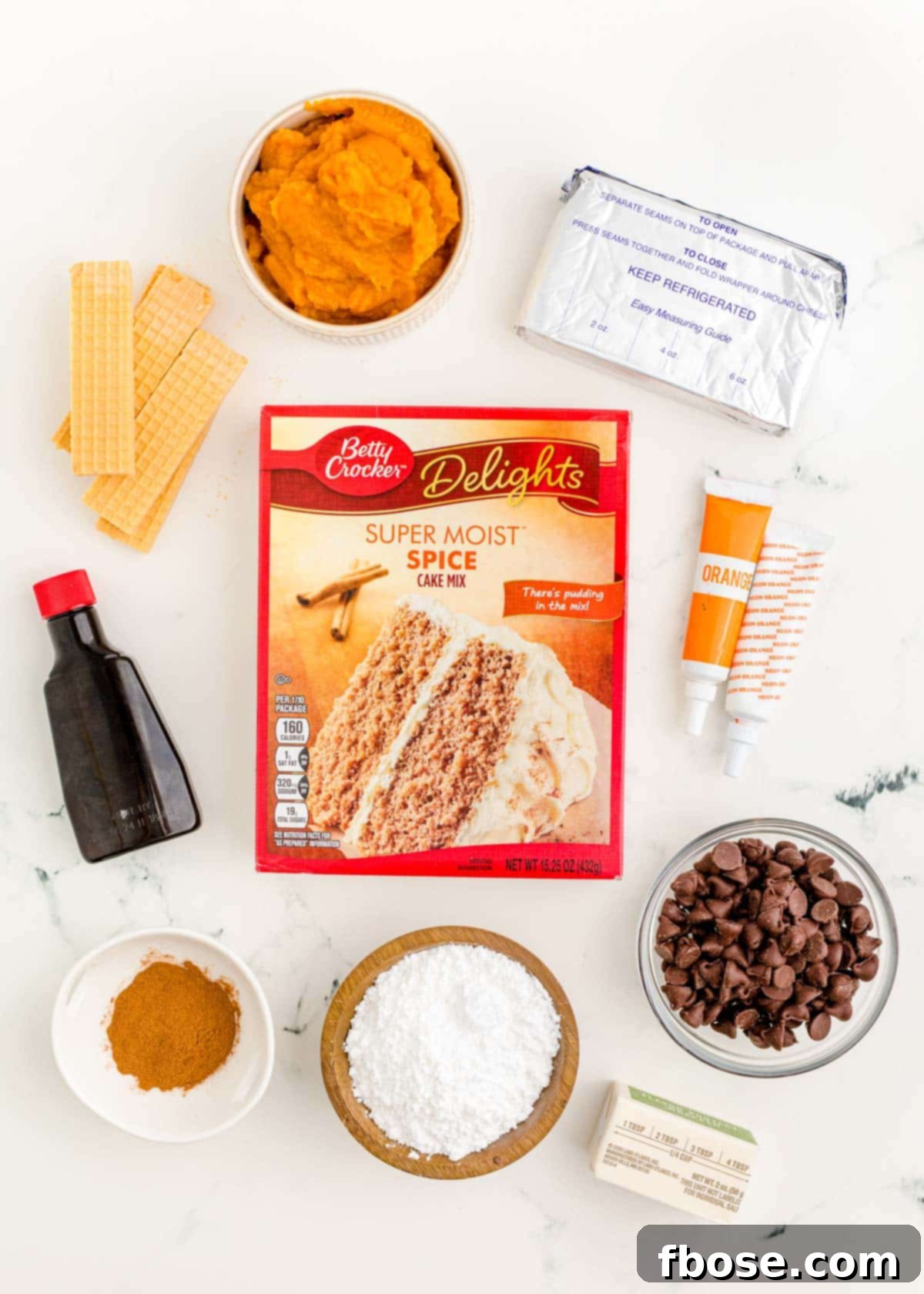 A vibrant flat lay shot displaying all the fresh and packaged ingredients needed to make mini pumpkin bundt cakes laid out on a clean white marble surface.