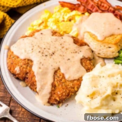 Country fried steak on a white plate with mashed potatoes, eggs, bacon, and a biscuit.