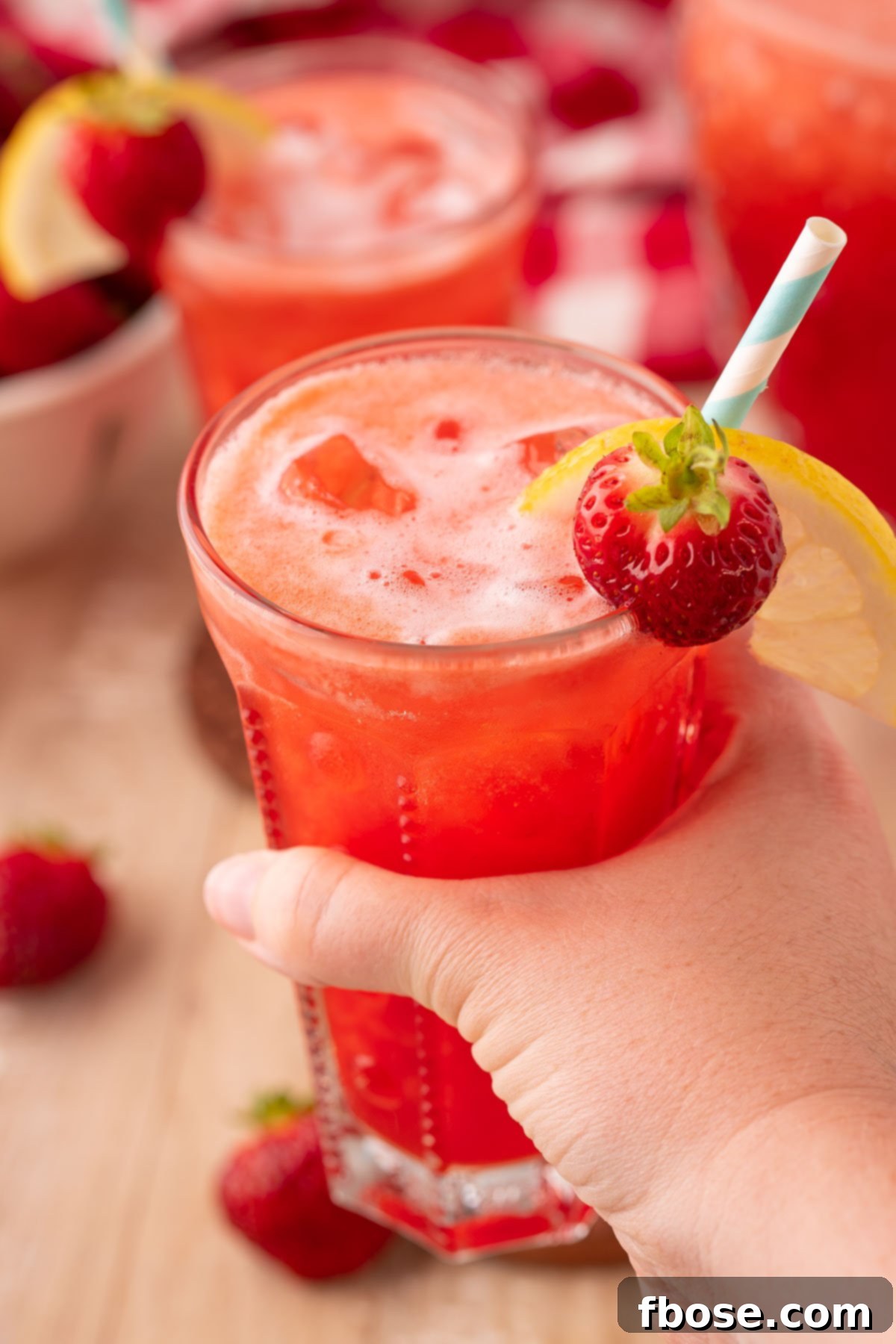 A woman's hand gently holds a glass of sparkling strawberry lemonade, beautifully garnished with a fresh strawberry and lemon slice, against a soft-focused background, emphasizing its refreshing appeal.