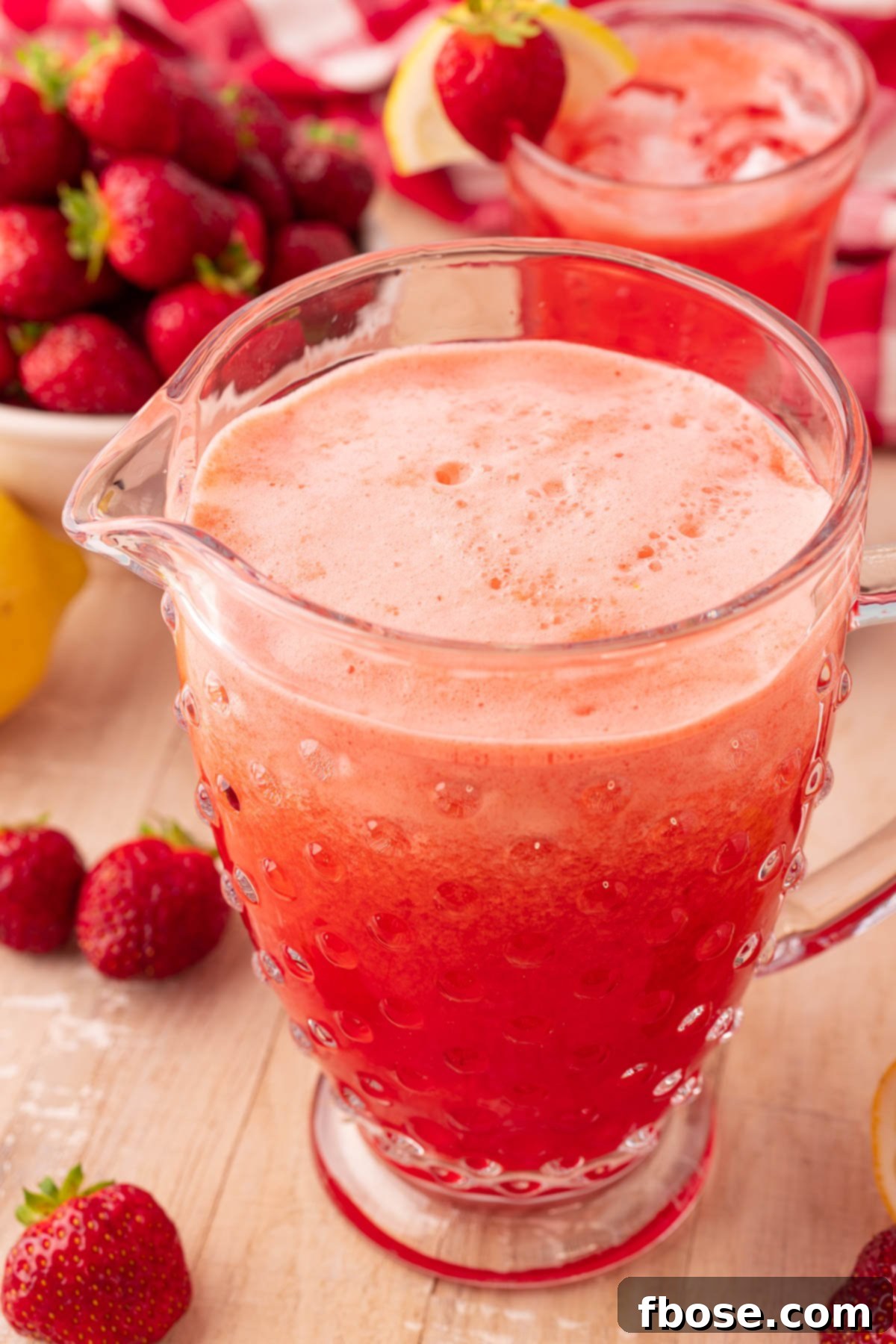 A large glass pitcher filled with homemade strawberry lemonade, garnished with fresh lemon slices and whole strawberries, sitting on a light wooden table.
