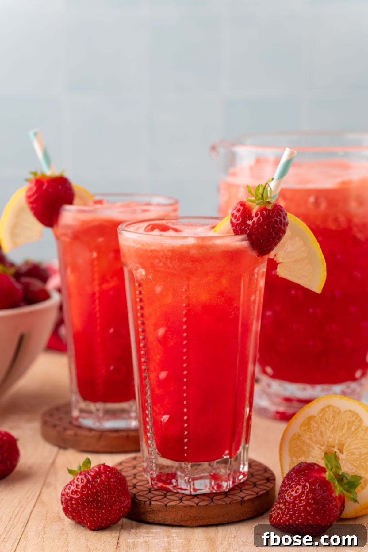 Two glasses of sparkling strawberry lemonade, beautifully garnished with fresh strawberries and lemon slices, resting on wooden coasters on a wooden table, ready to be enjoyed.