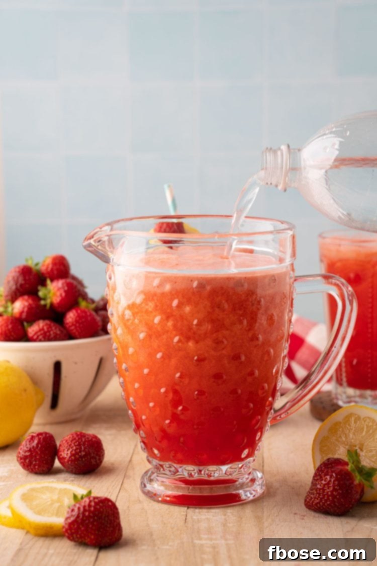 Fizzy club soda being gently poured into a large pitcher already containing the strawberry lemonade concentrate, ready to be stirred and served.