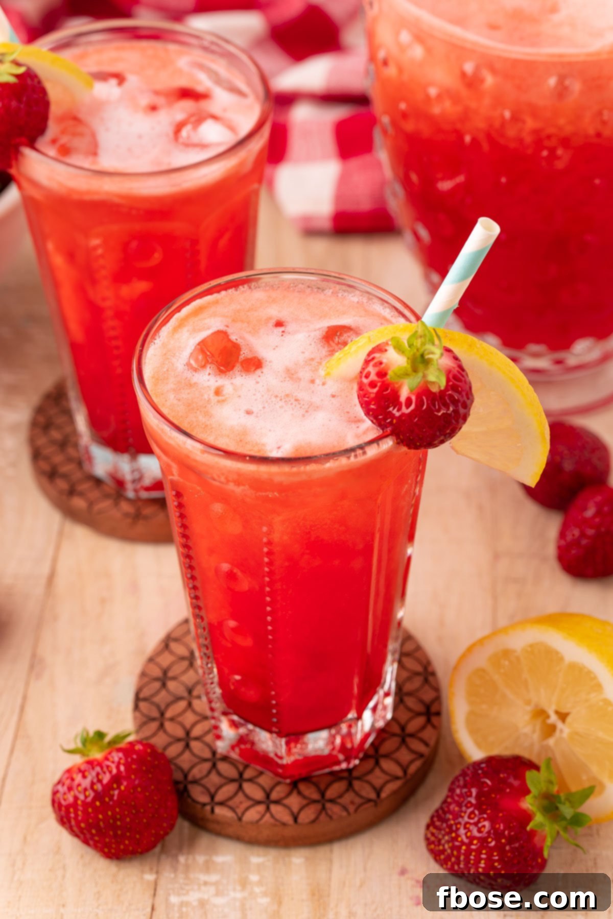 Multiple glasses of vibrant pink strawberry lemonade, adorned with fresh fruit garnishes, sitting on individual wooden coasters on a light-colored wooden table, inviting refreshment.