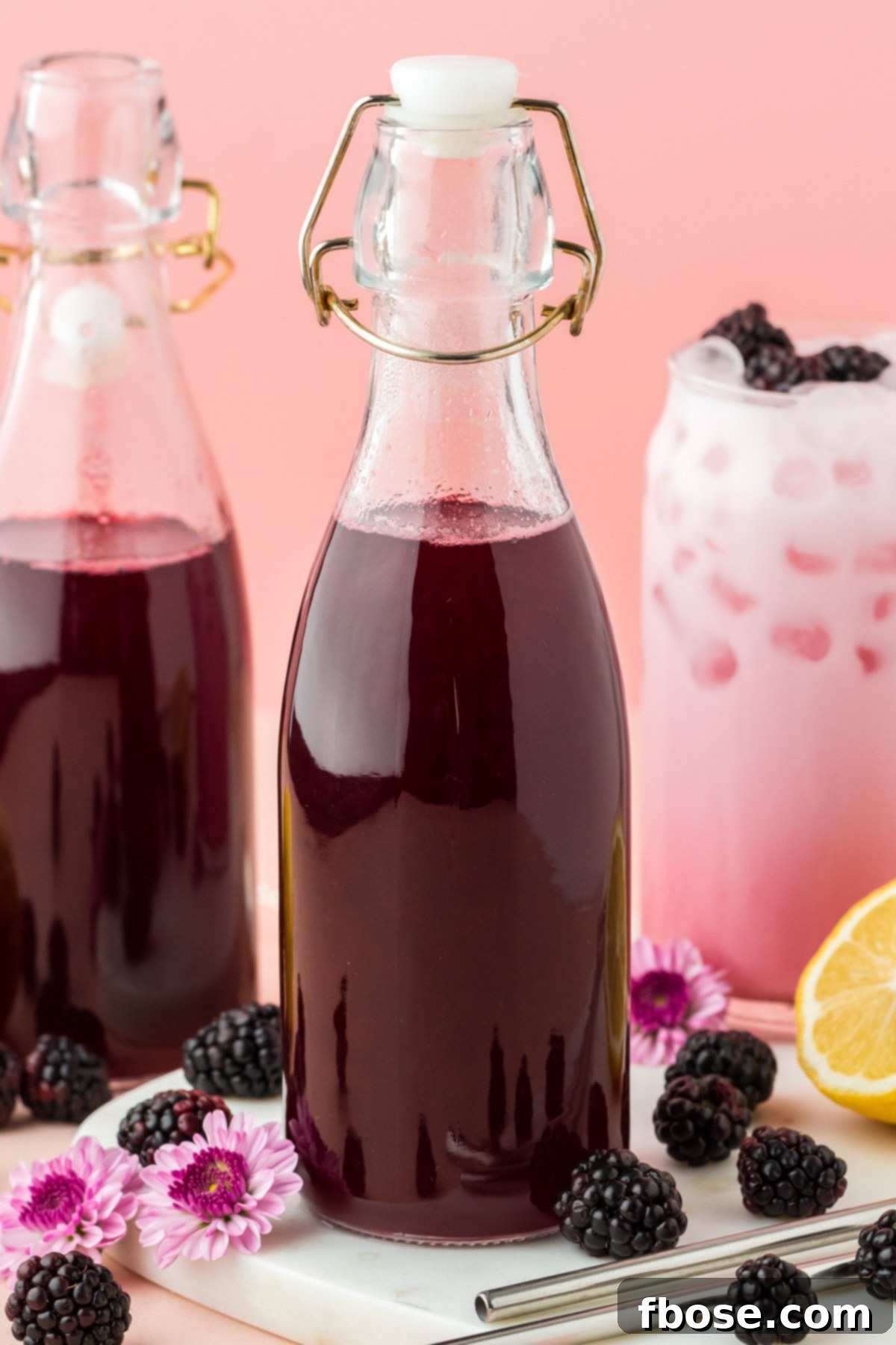 Close up photo of two elegant glass bottles of homemade blackberry simple syrup alongside a glass of blackberry cream soda, garnished with fresh blackberries.