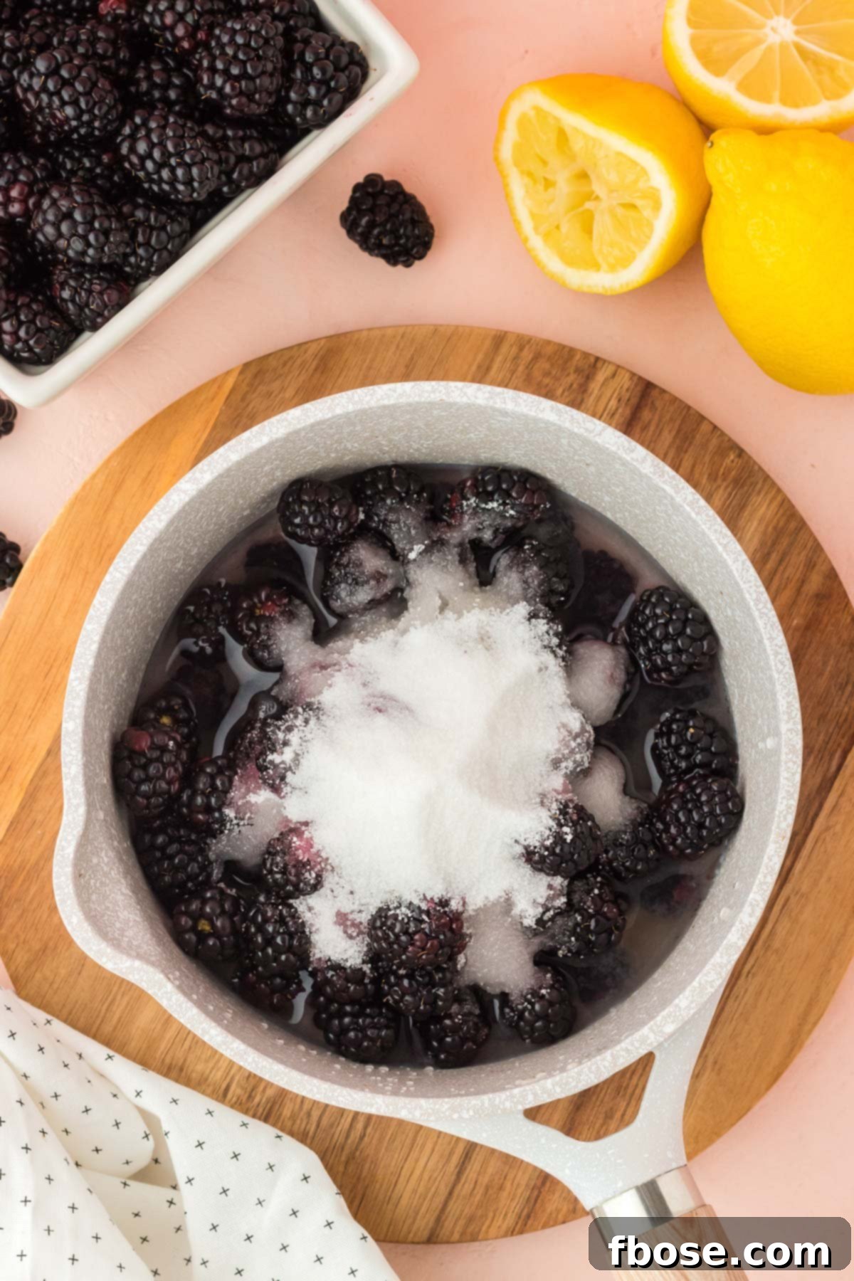 Overhead shot of a saucepan containing fresh blackberries, white granulated sugar, and clear water, poised on a stovetop, ready for cooking.
