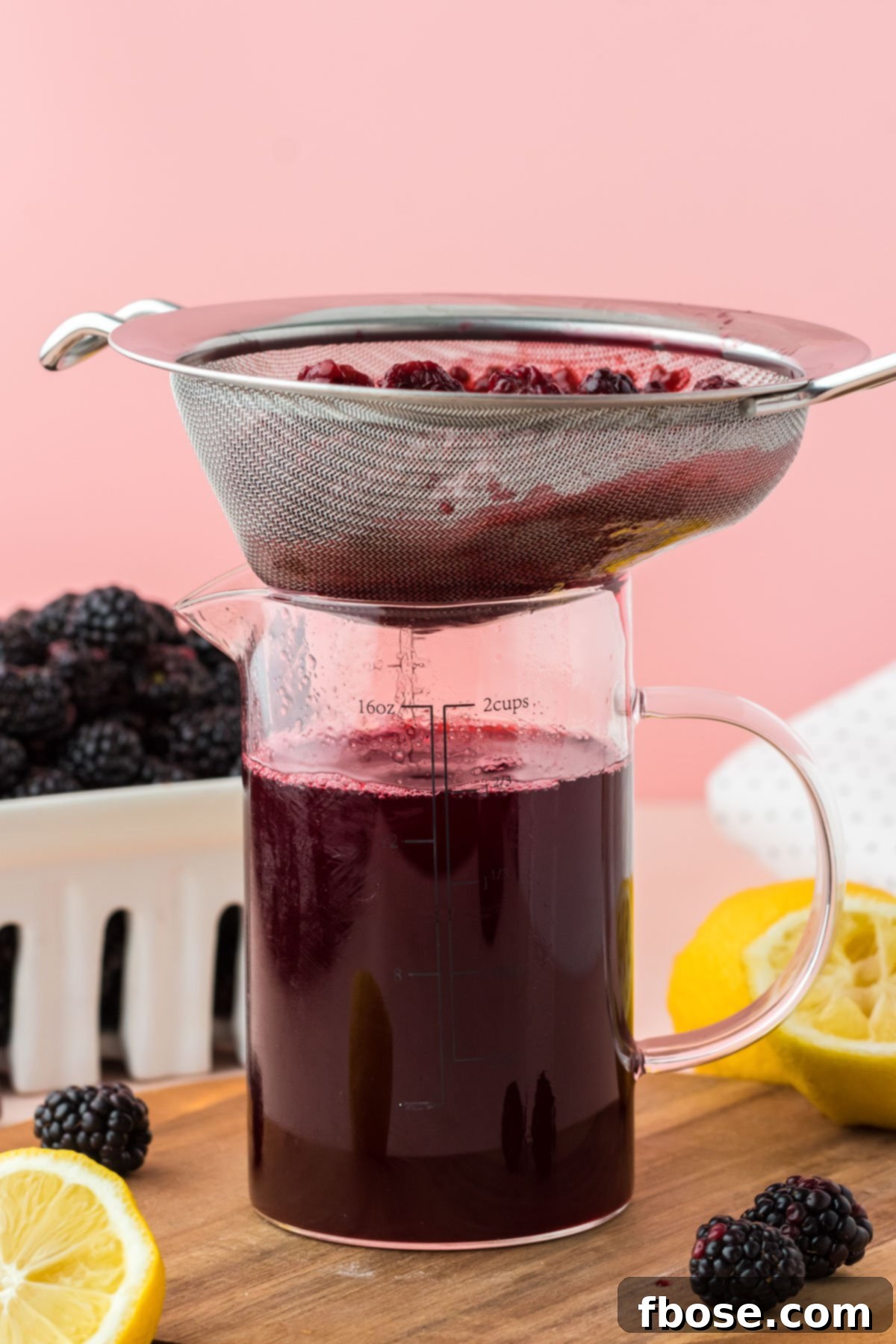 A fine mesh sieve actively straining homemade blackberry simple syrup into a clear glass measuring cup, separating the smooth liquid from the berry pulp.