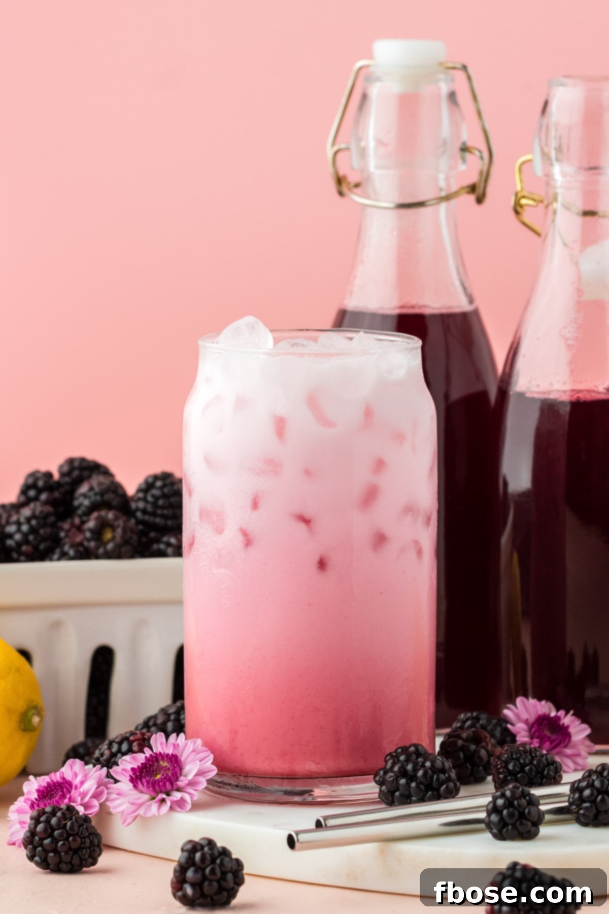A refreshing glass of blackberry Italian cream soda sits on a white serving board, with a bottle of the homemade syrup and fresh blackberries blurred in the background, hinting at its delicious origin.
