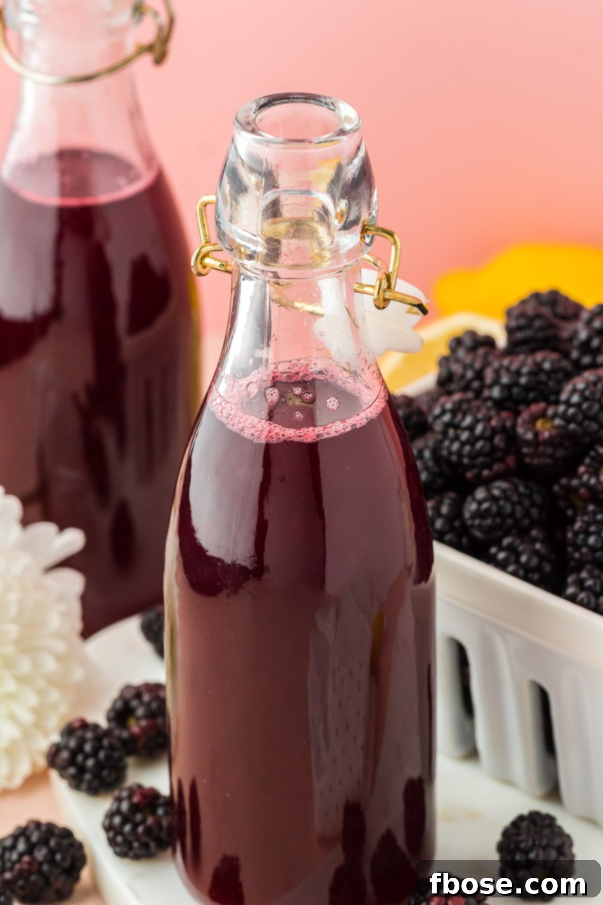 Close-up shot of a single bottle of blackberry simple syrup in the foreground, with another bottle and a scattering of fresh blackberries artfully blurred in the background.