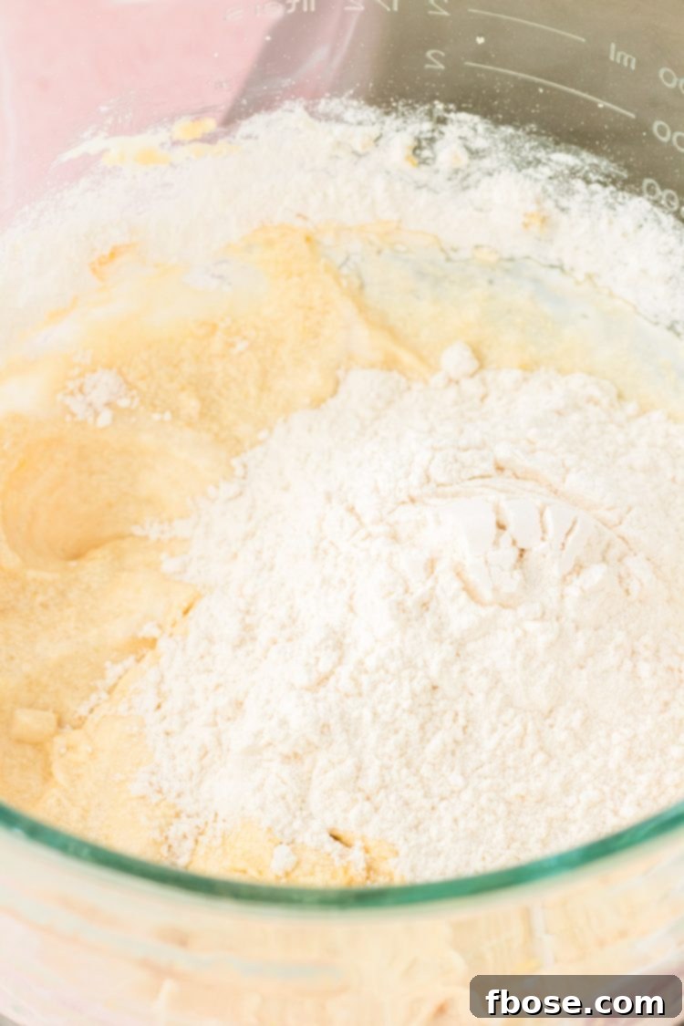 A spoonful of dry ingredients being added into a bowl of wet cake batter, illustrating the alternating mixing process.