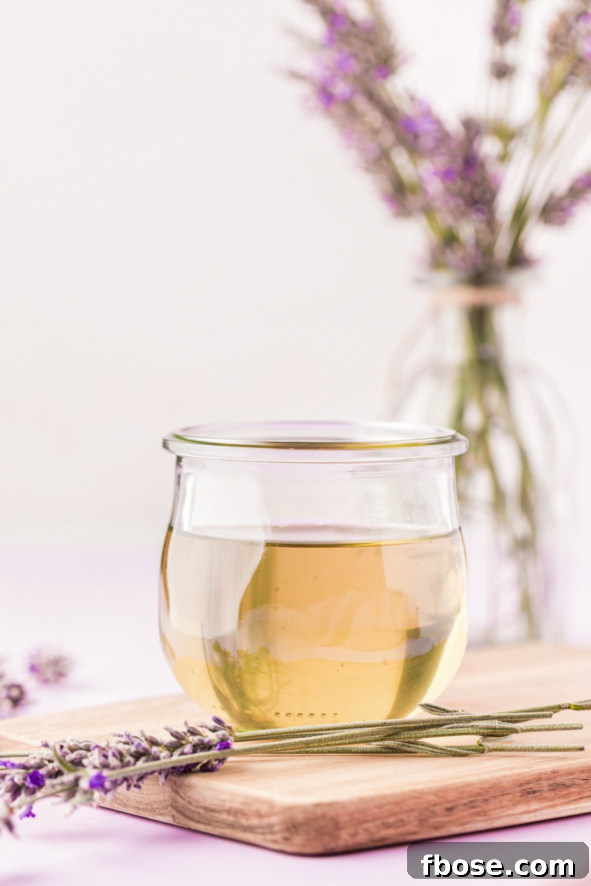 A small jar of lavender simple syrup on a wooden board with fresh lavender sprigs surrounding it.