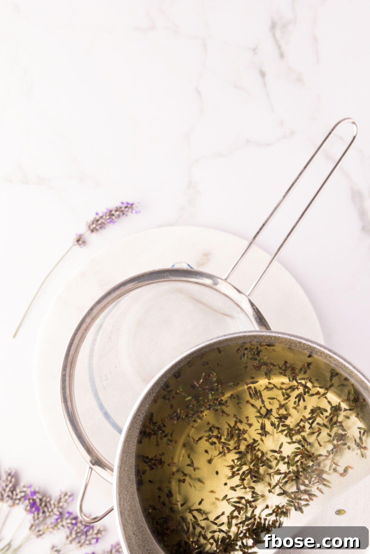 Lavender simple syrup being strained into a glass measuring cup through a fine-mesh sieve.