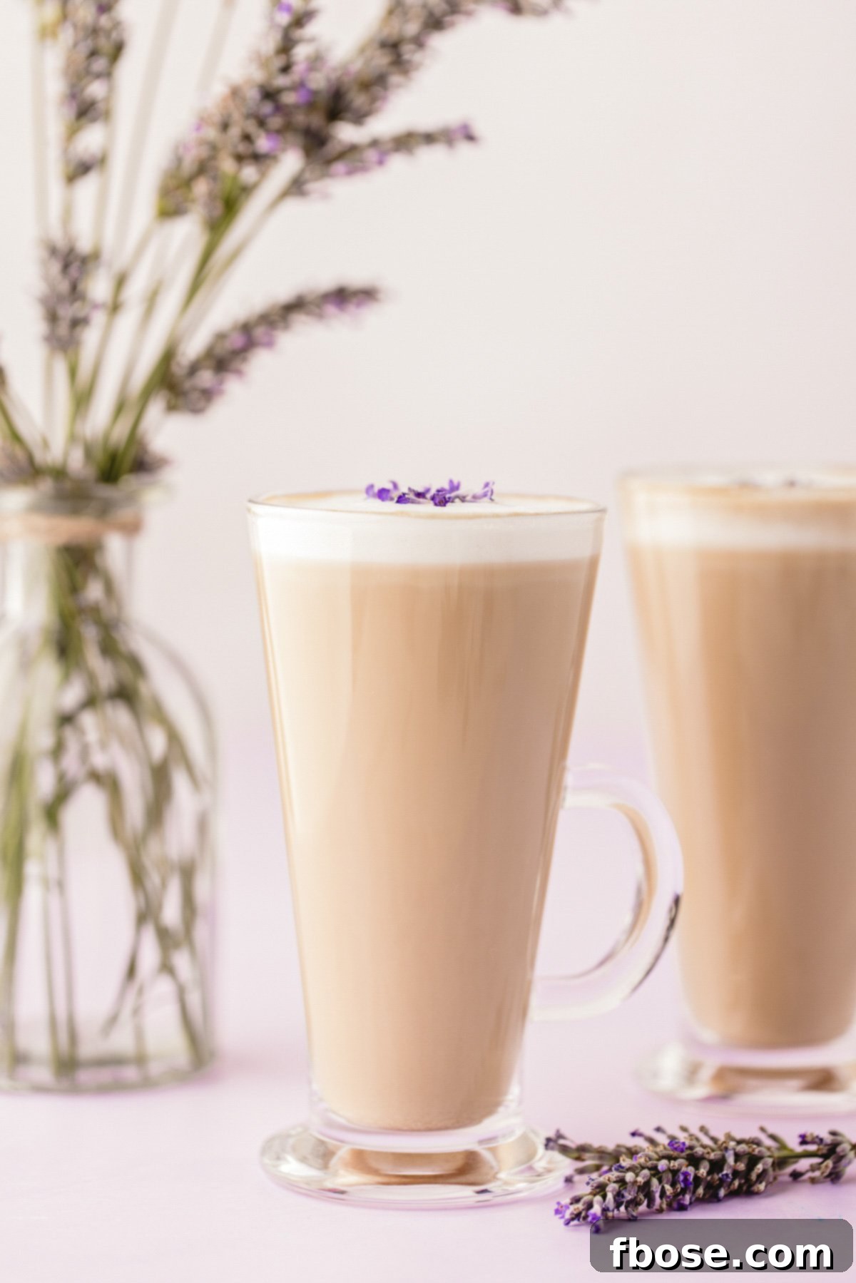 A tapered glass mug with a perfectly frothed lavender latte, another inviting mug gently blurred in the background, set against a warm, rustic backdrop.
