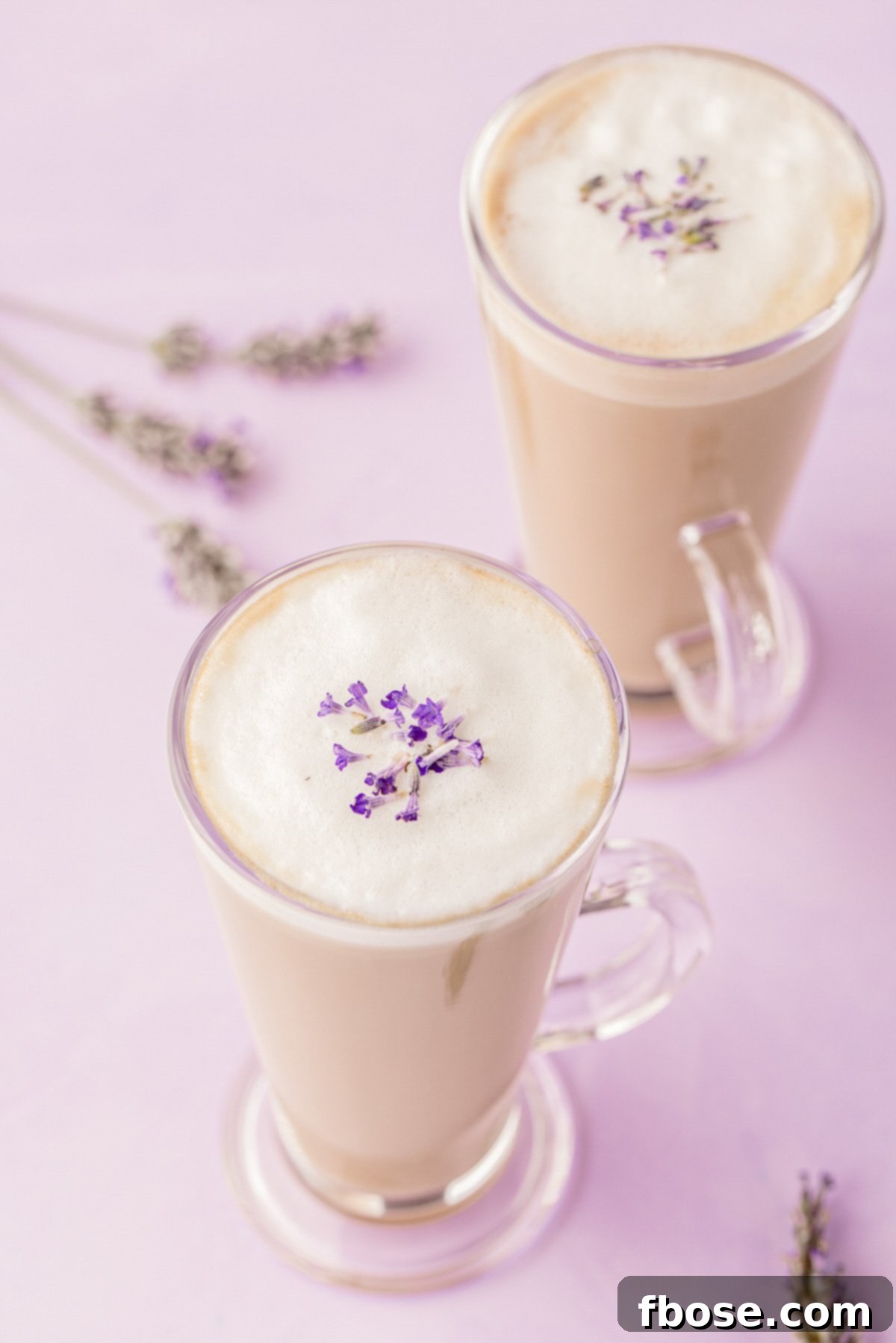 Two aesthetically pleasing lavender lattes presented in clear glass mugs, resting on a smooth purple surface, highlighting their inviting color and delicate foam.