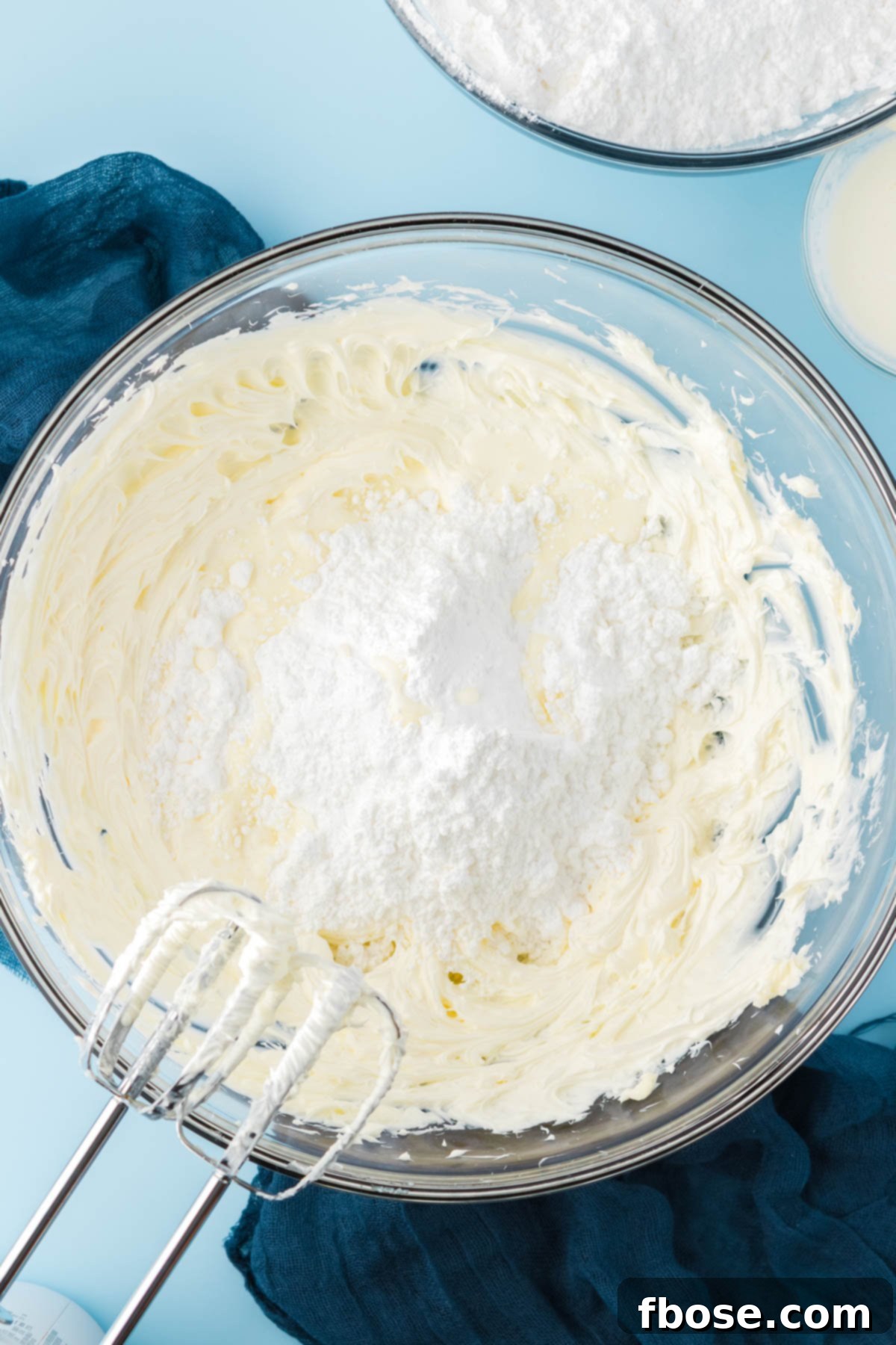 Powdered sugar being added to a glass bowl with creamed butter to make frosting.