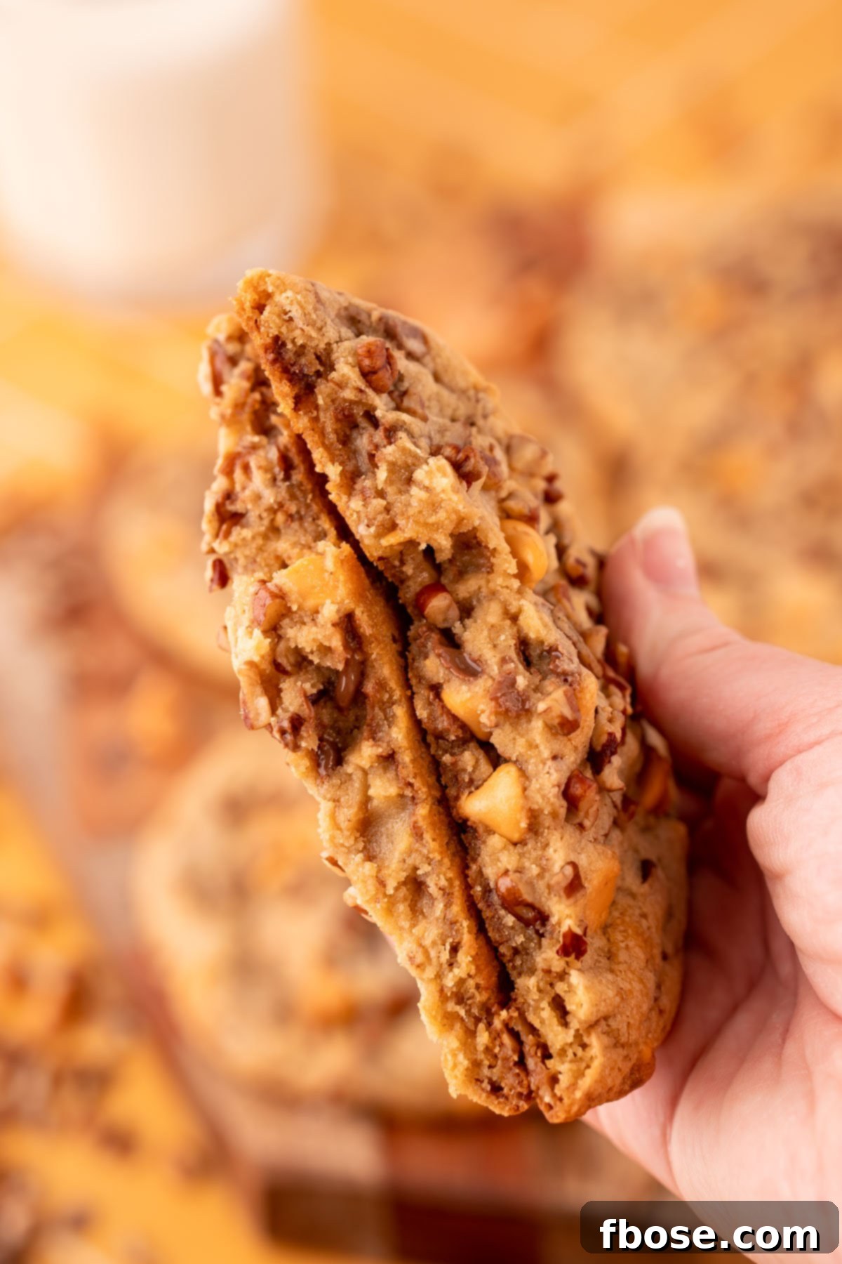 A woman's hand holds a large butter pecan cookie broken in half, revealing its soft, chewy center filled with pecans and chips.