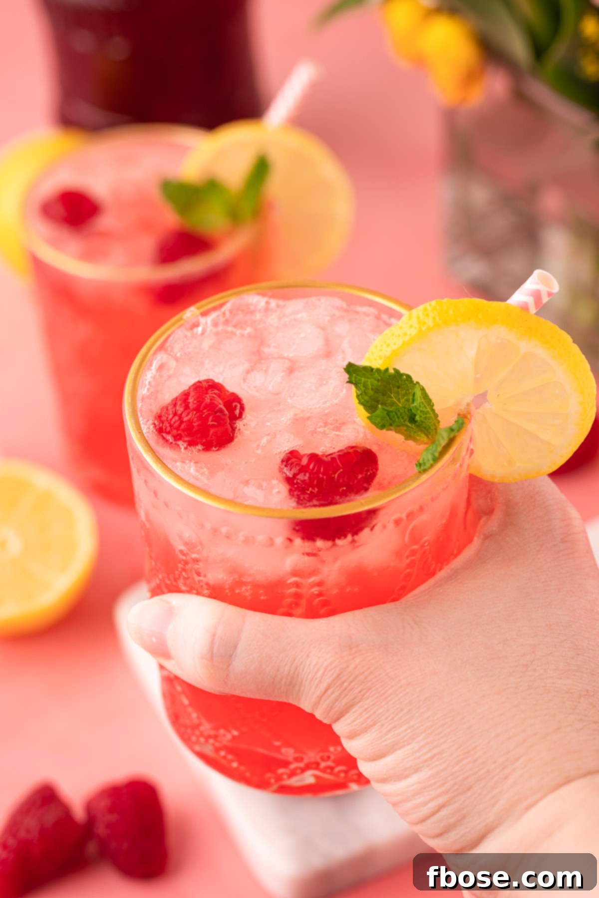 A woman's hand elegantly holds a garnished Raspberry Gin Daisy cocktail, presenting it to the camera with a inviting gesture. The vibrant colors of the drink and its garnishes are prominent.