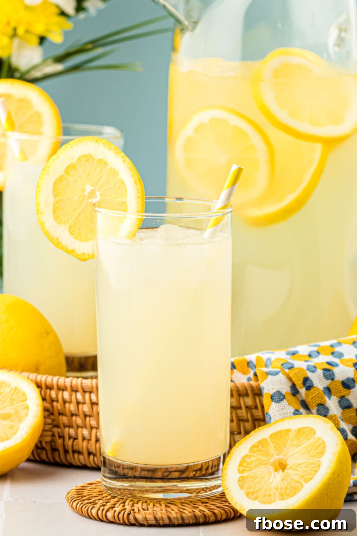 Homemade Roasted Lemonade in a clear glass with ice and a lemon slice, with a pitcher of lemonade in the background.
