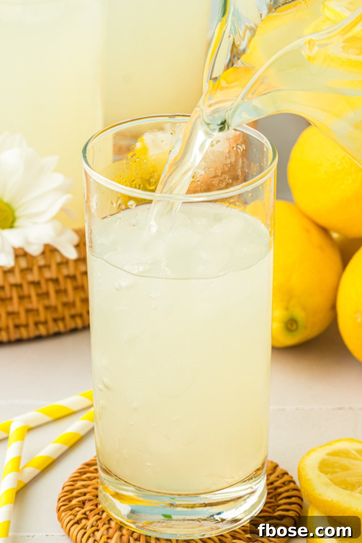 A stream of golden lemonade being poured from a pitcher into a tall glass filled with ice, ready to drink.