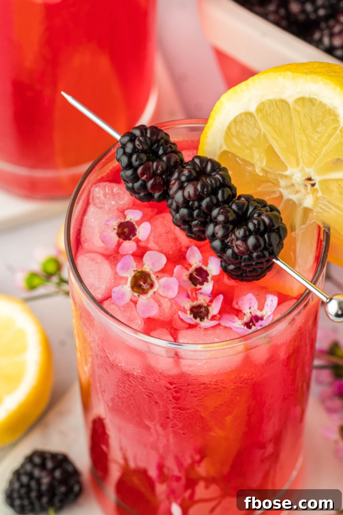 Delicious Homemade Blackberry Lemonade 3 Fresh blackberry lemonade in a clear glass, garnished with a slice of lemon, whole blackberries, and delicate flowers, presented beautifully on a clean surface.