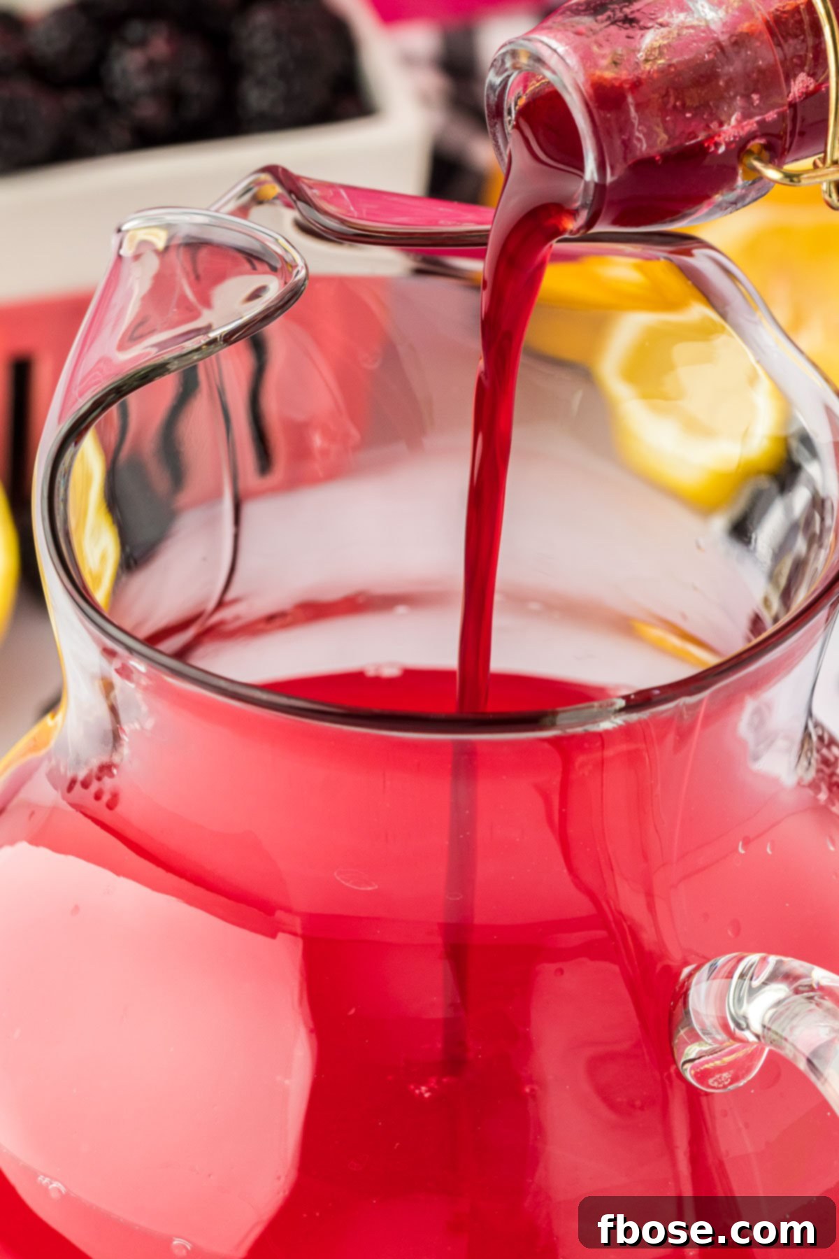 Delicious Homemade Blackberry Lemonade 8 Rich blackberry simple syrup being poured from a small saucepan into a large, clear glass pitcher, ready to mix with lemon juice and water.