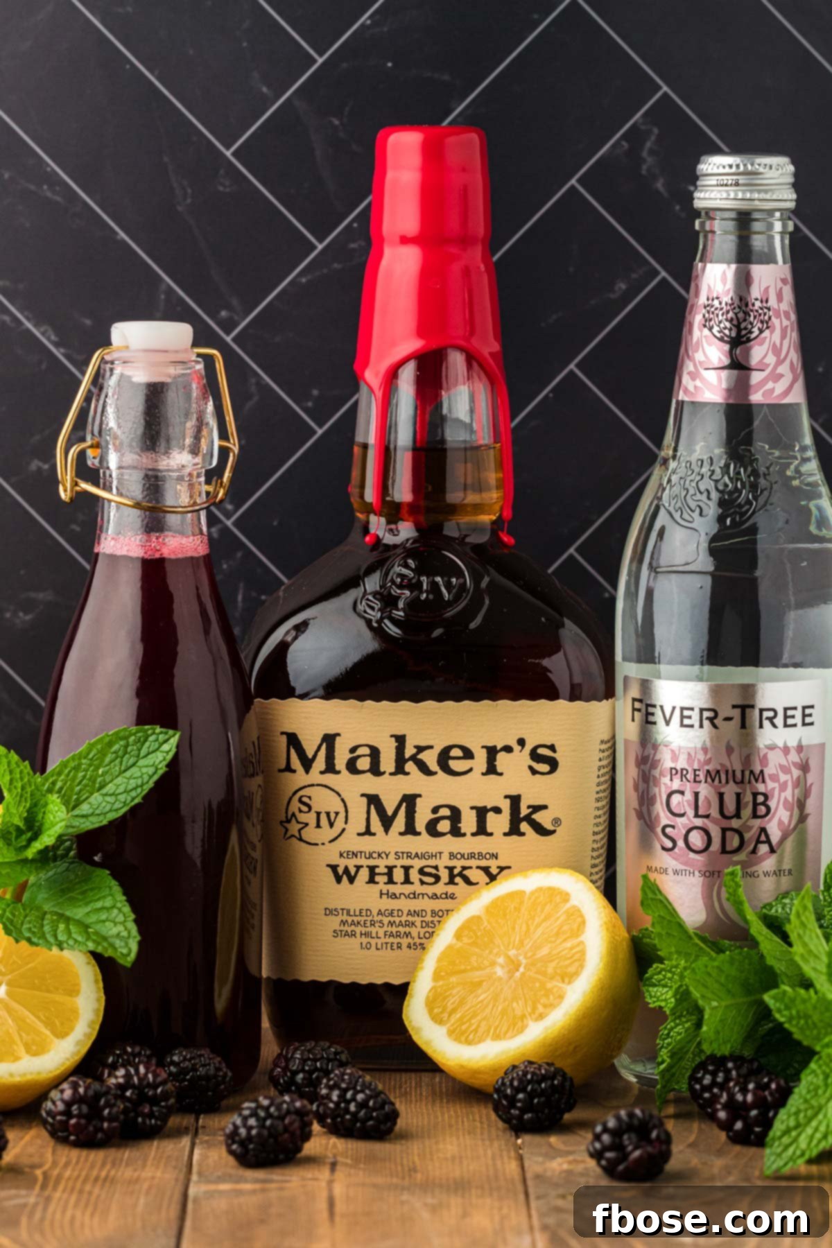 Ingredients to make a bourbon smash (blackberry) on a wooden table, including fresh mint, lemon, blackberries, and bourbon.