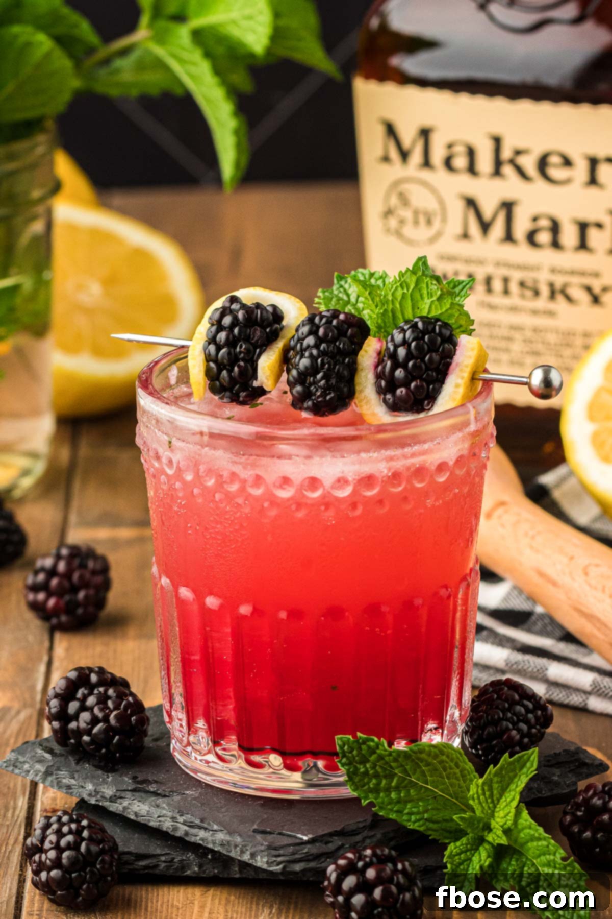 A glass with a bourbon smash (blackberry flavored) on a black coaster, garnished with a lemon slice and blackberries.