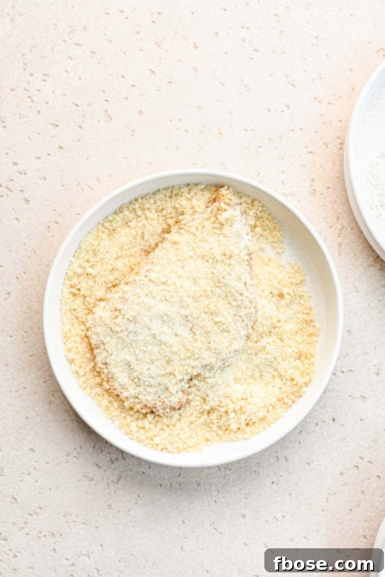 A chicken breast being generously coated with panko bread crumbs in a white mixing bowl.