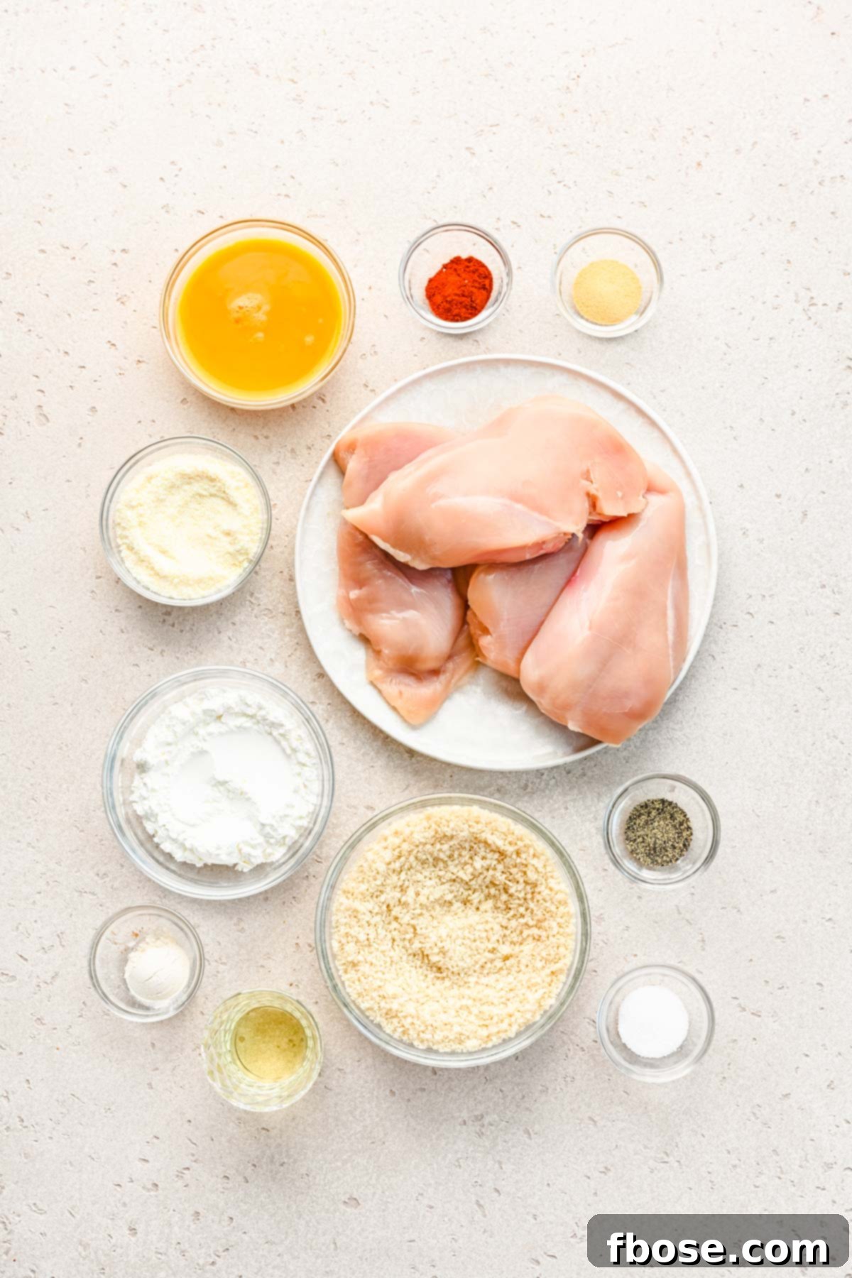 Overhead shot of various ingredients laid out on a kitchen counter: chicken breasts, bowls of cornstarch, spices, eggs, panko breadcrumbs, and Parmesan cheese.
