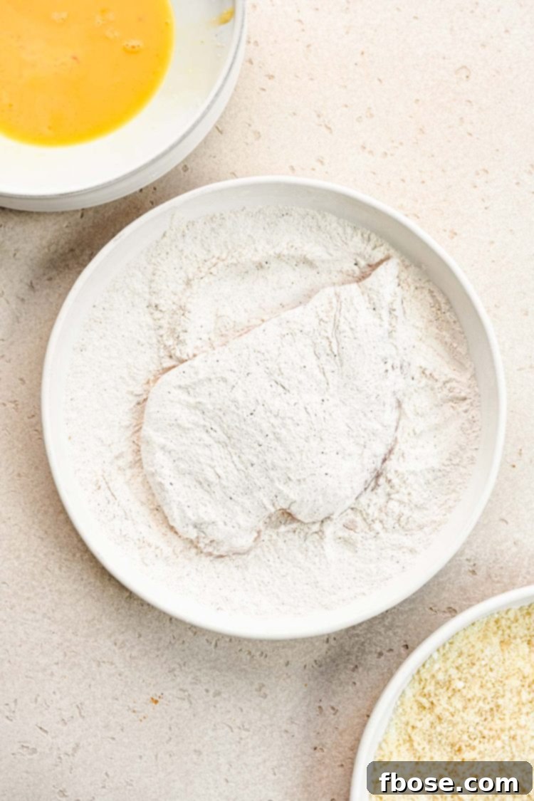 A raw chicken breast being coated in a dry flour mixture in a shallow dish.