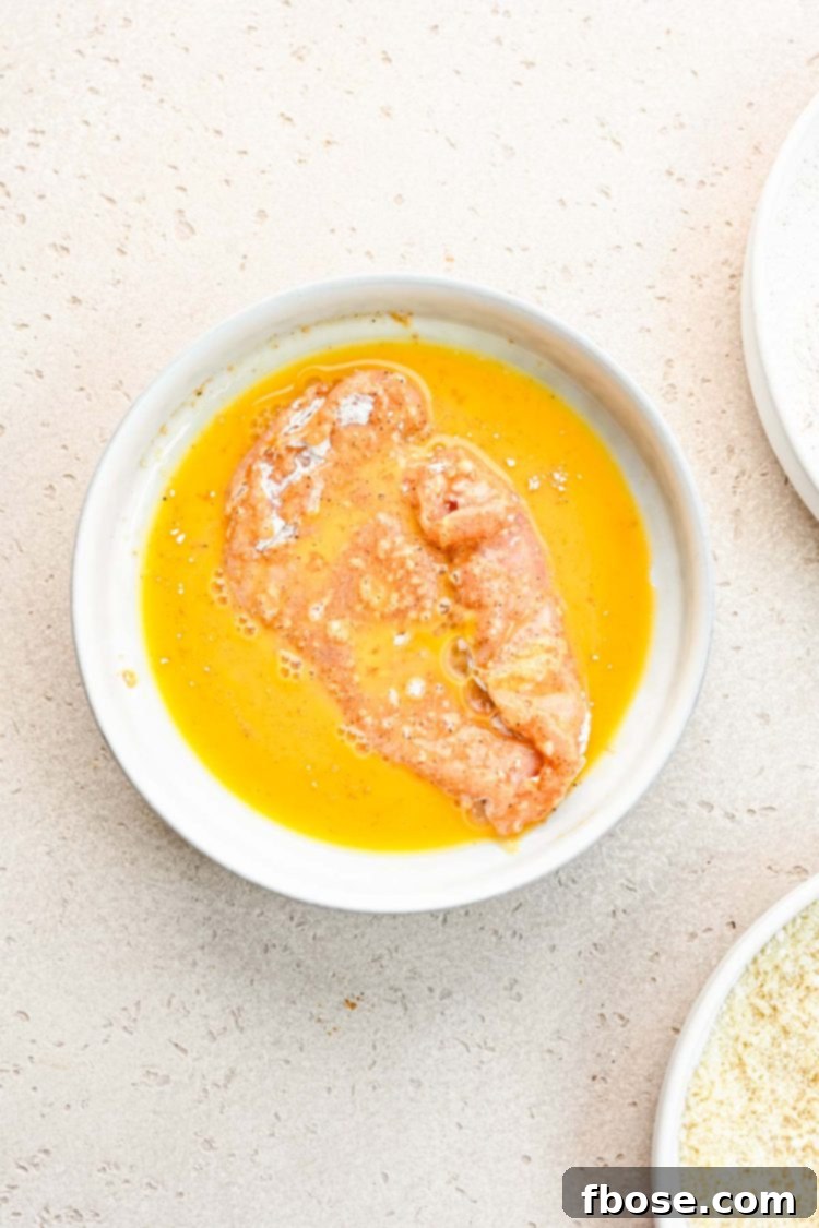A chicken breast, already coated in flour, being dipped into beaten egg in a bowl.