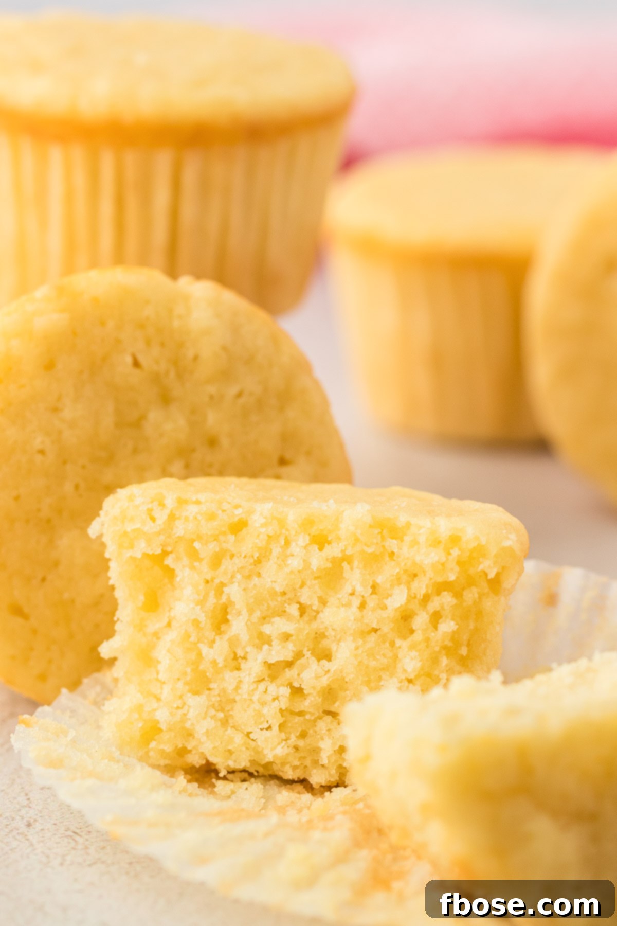 A perfectly baked plain yellow cupcake, sliced in half to reveal its soft, tender crumb. Other cupcakes are blurred in the background.
