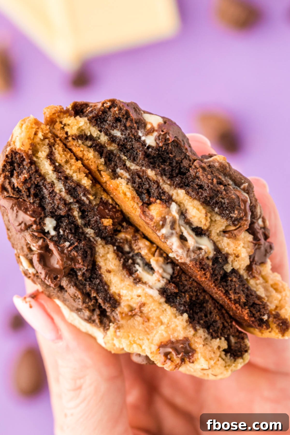 A woman's hand delicately holding a Kitchen Sink Cookie, cut in half to showcase its impressive array of internal ingredients and marbled texture.