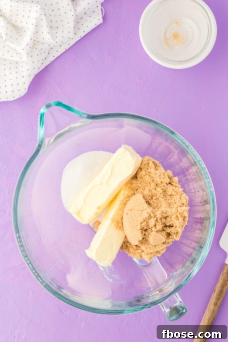 Softened butter and sugars being creamed together in a clear glass mixing bowl, forming a pale, fluffy mixture.