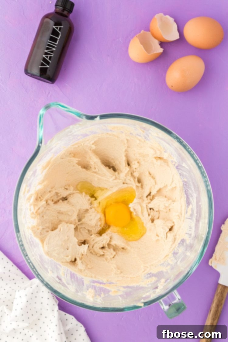 A large egg being cracked and added to the creamed butter and sugar mixture in a glass bowl, illustrating the next step in cookie dough preparation.