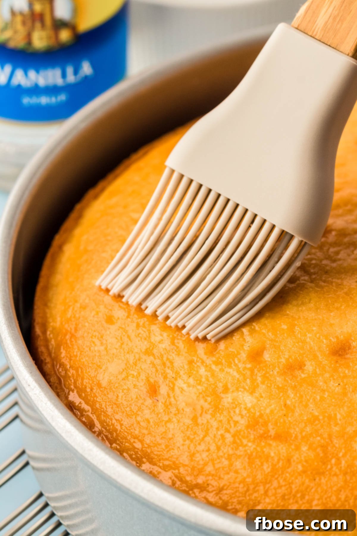 A pastry brush glides across the top of a freshly baked white vanilla cake, generously applying vanilla simple syrup for added moisture and flavor.