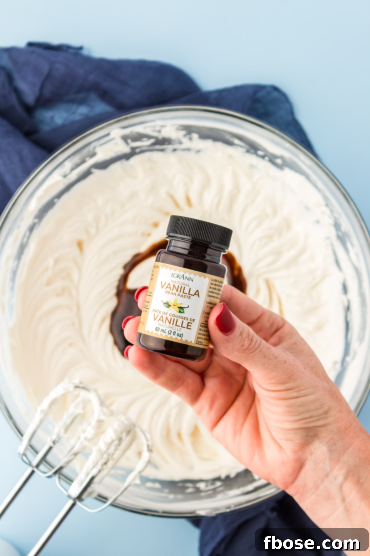 A woman's hand holds a bottle of vanilla bean paste, ready to add its rich essence to a bowl of creamy vanilla frosting.