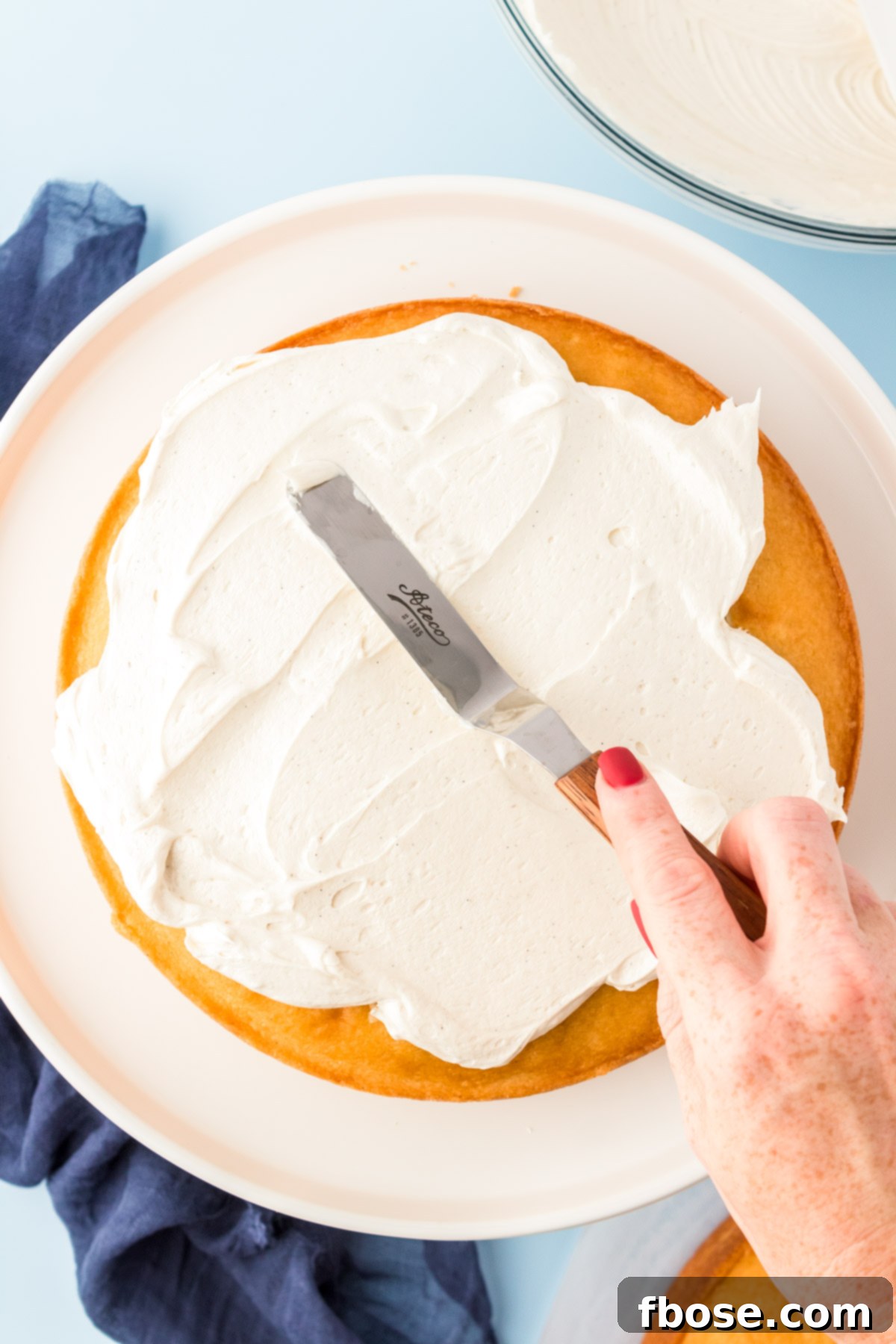 A woman's hand expertly spreads a layer of creamy frosting over a cake using an offset spatula, creating a smooth, even coating.