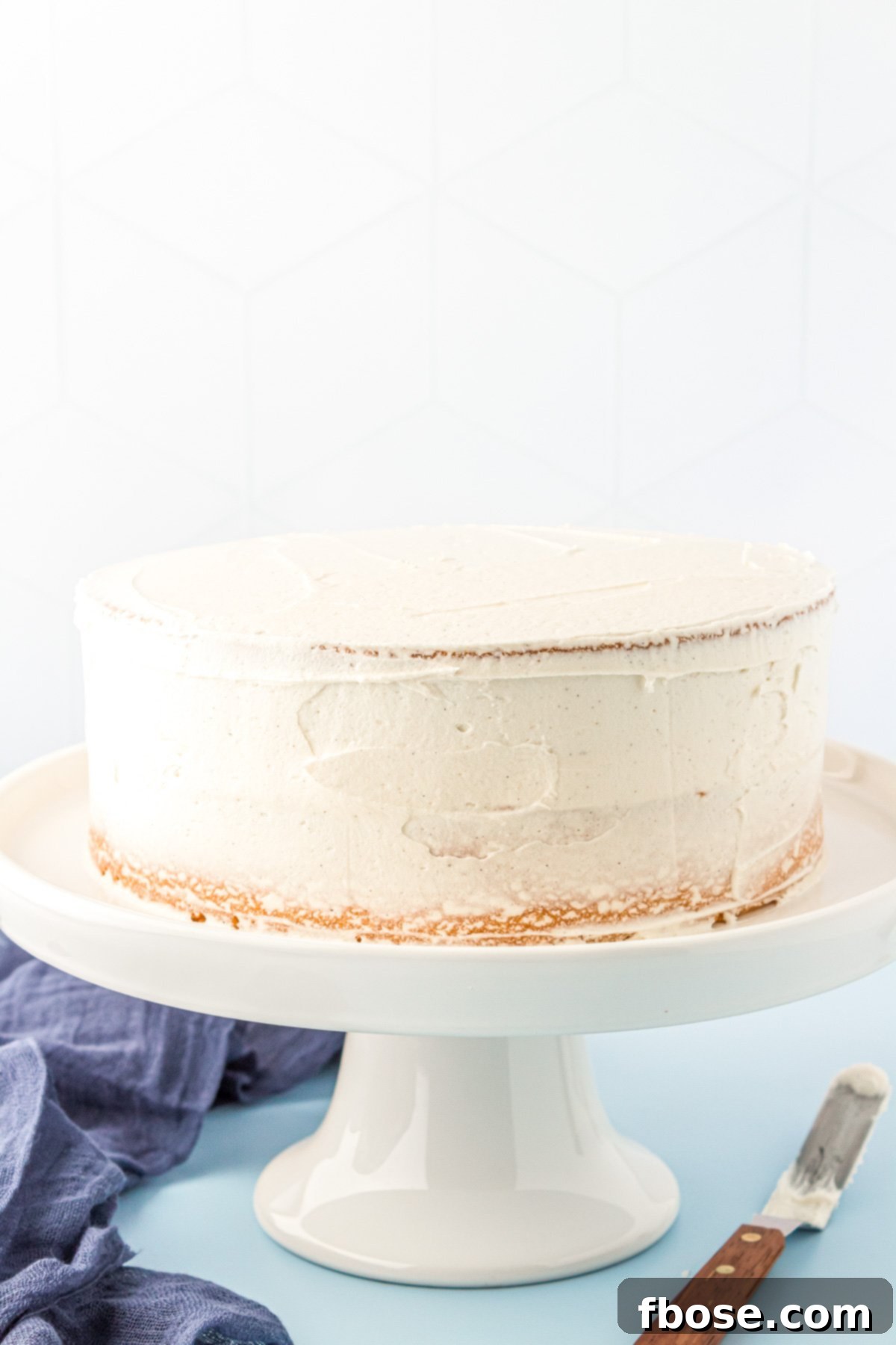 A beautifully crumb-coated vanilla cake rests on a pristine white cake stand, its thin layer of vanilla frosting ready for its final flourish.