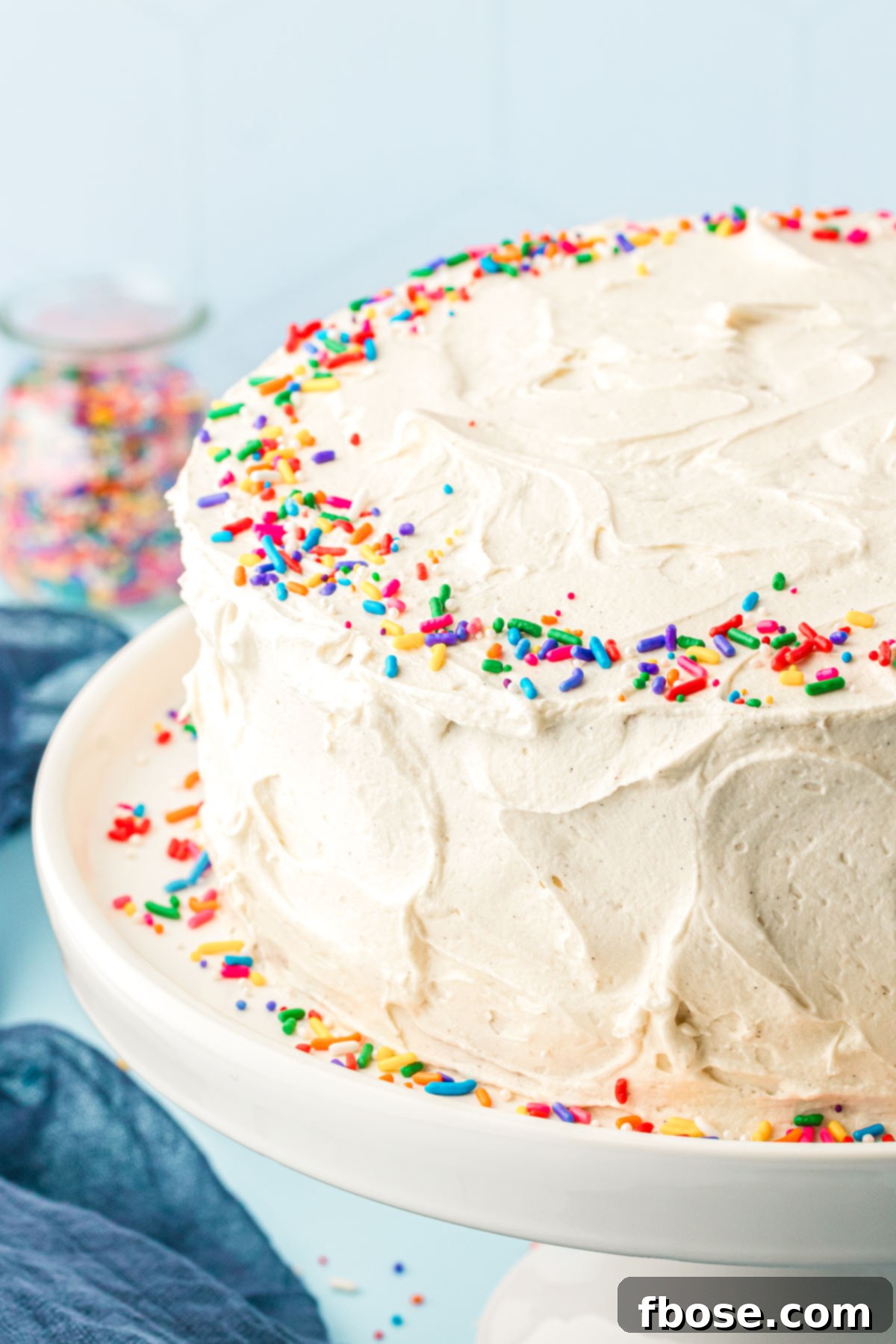 A celebratory vanilla frosted cake adorned with vibrant rainbow sprinkles, elegantly displayed on a white cake stand, ready to be enjoyed.