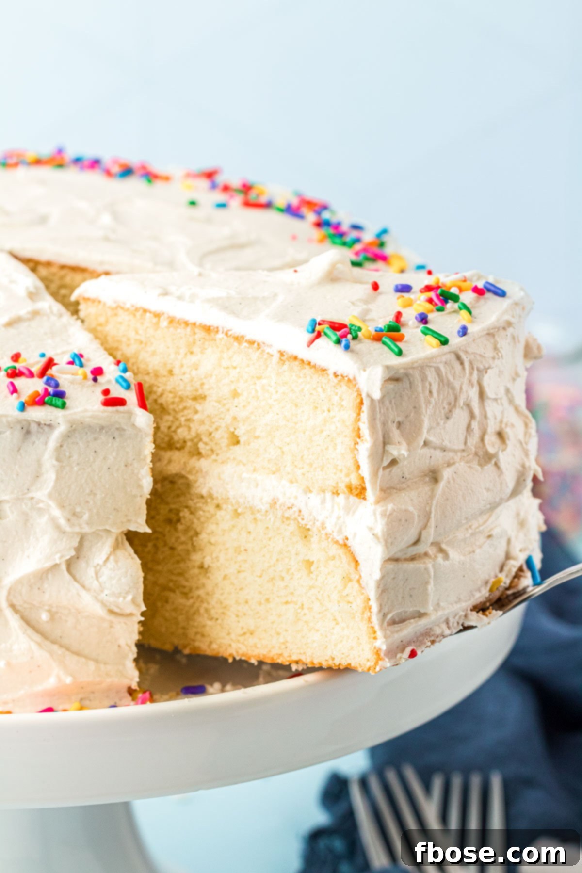 A cake server carefully lifts a perfect slice of vanilla cake from a white cake stand, highlighting the moist layers and creamy frosting.