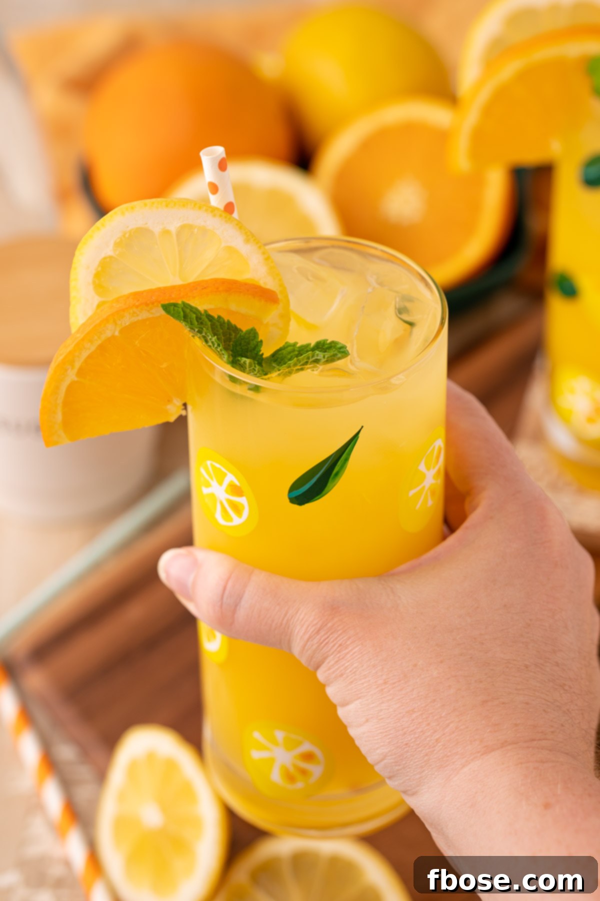 Sunkissed Orangeade 7 A woman's hand holding a clear glass of homemade orangeade, with an orange slice and mint garnish, against a bright background.