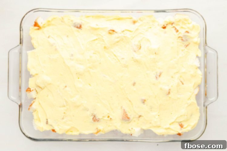 Smooth vanilla pudding mixture being spread evenly over the second layer of angel food cake in a pan, creating a creamy top layer.
