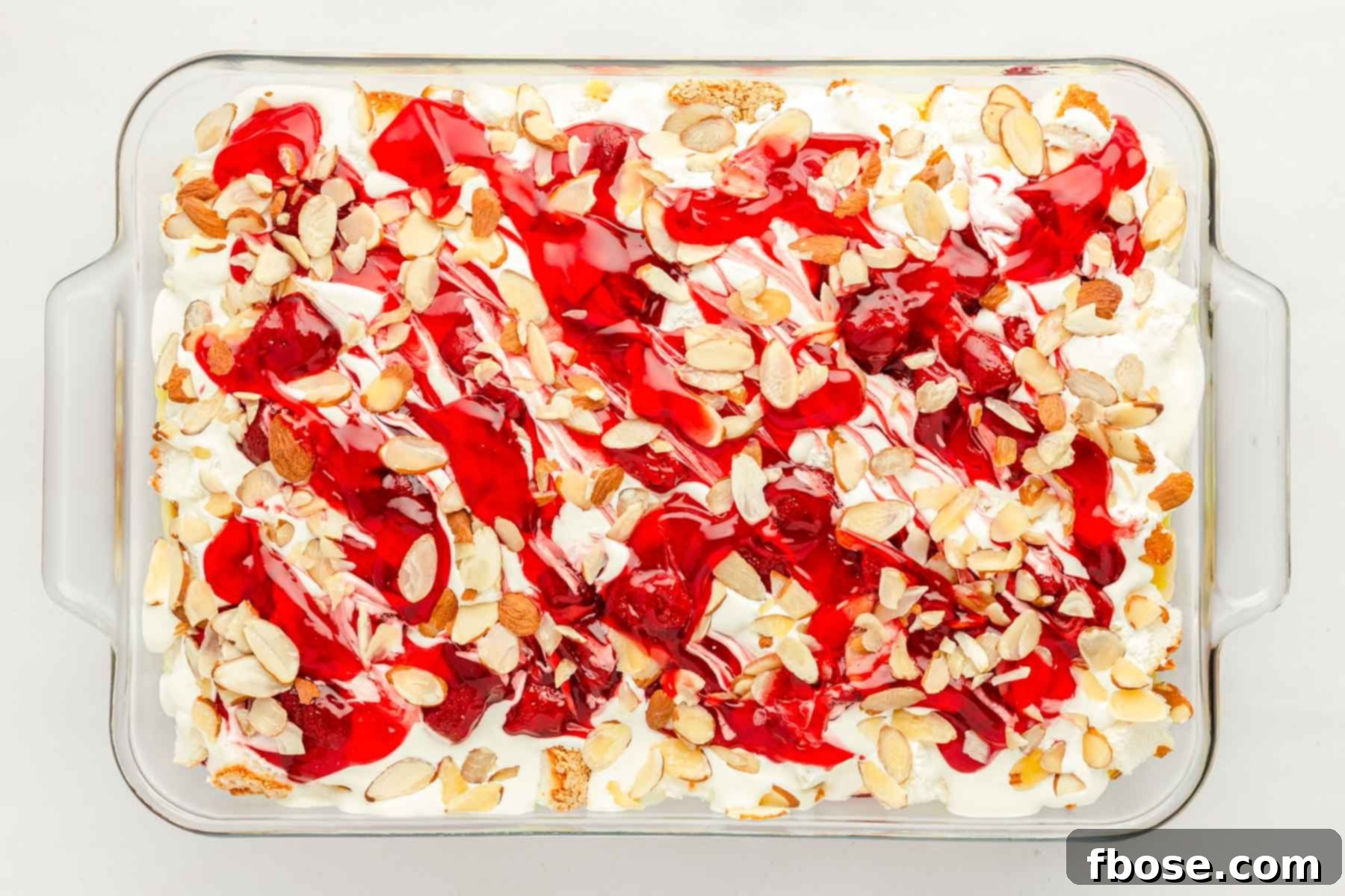 Overhead photo of a glass pan showcasing the fully assembled strawberry Heaven on Earth cake, topped with cool whip, strawberry swirls, and slivered almonds.