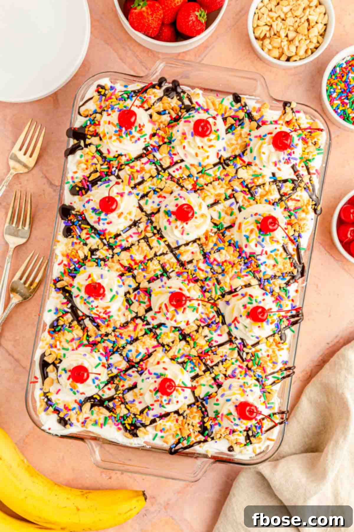 An inviting overhead photo of a completed no-bake banana split cake, garnished with chocolate syrup, nuts, sprinkles, and cherries, on a vibrant pink surface.