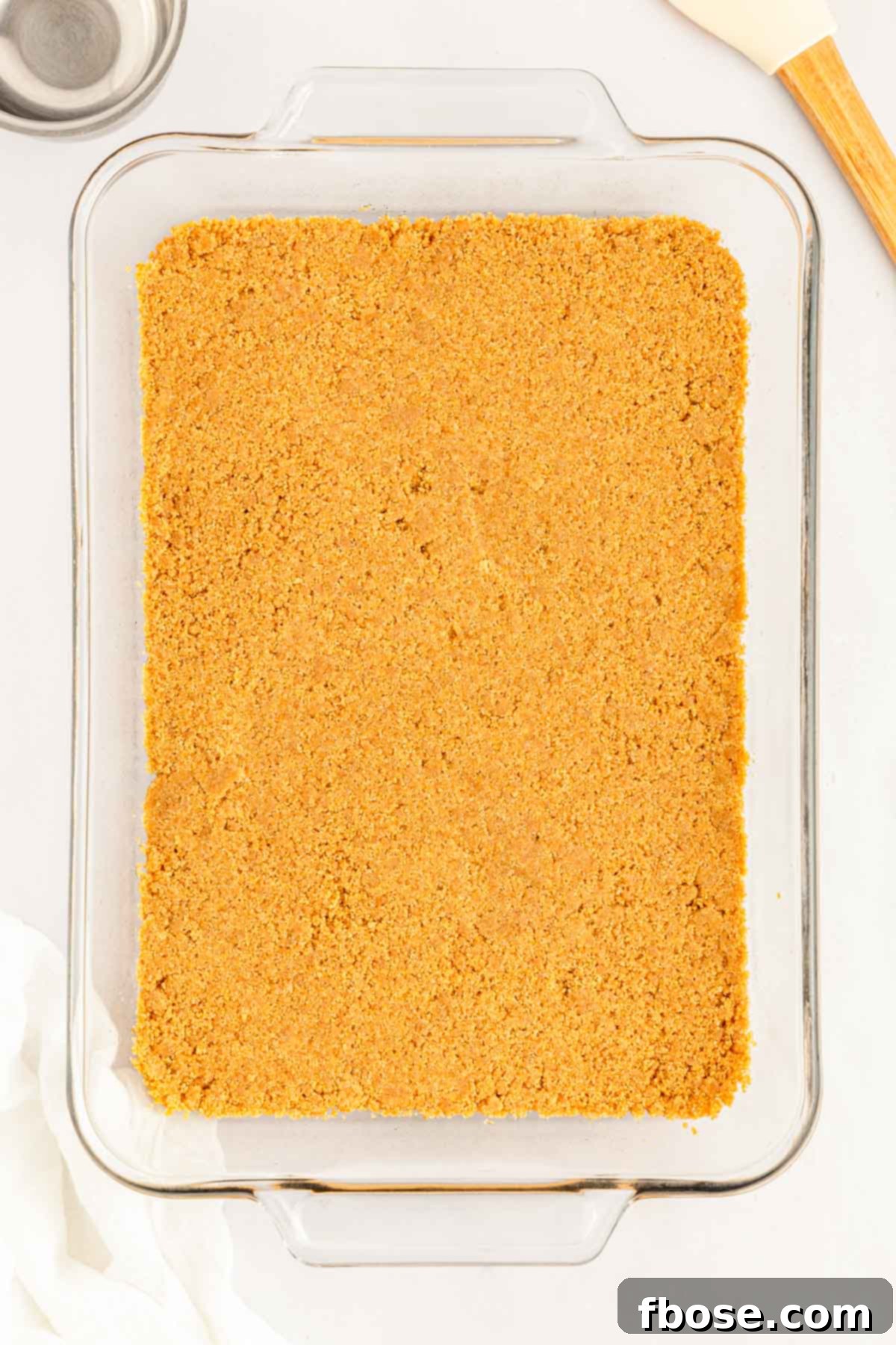 A perfectly pressed layer of golden graham cracker crumbs forming the crust in a clear glass baking pan.