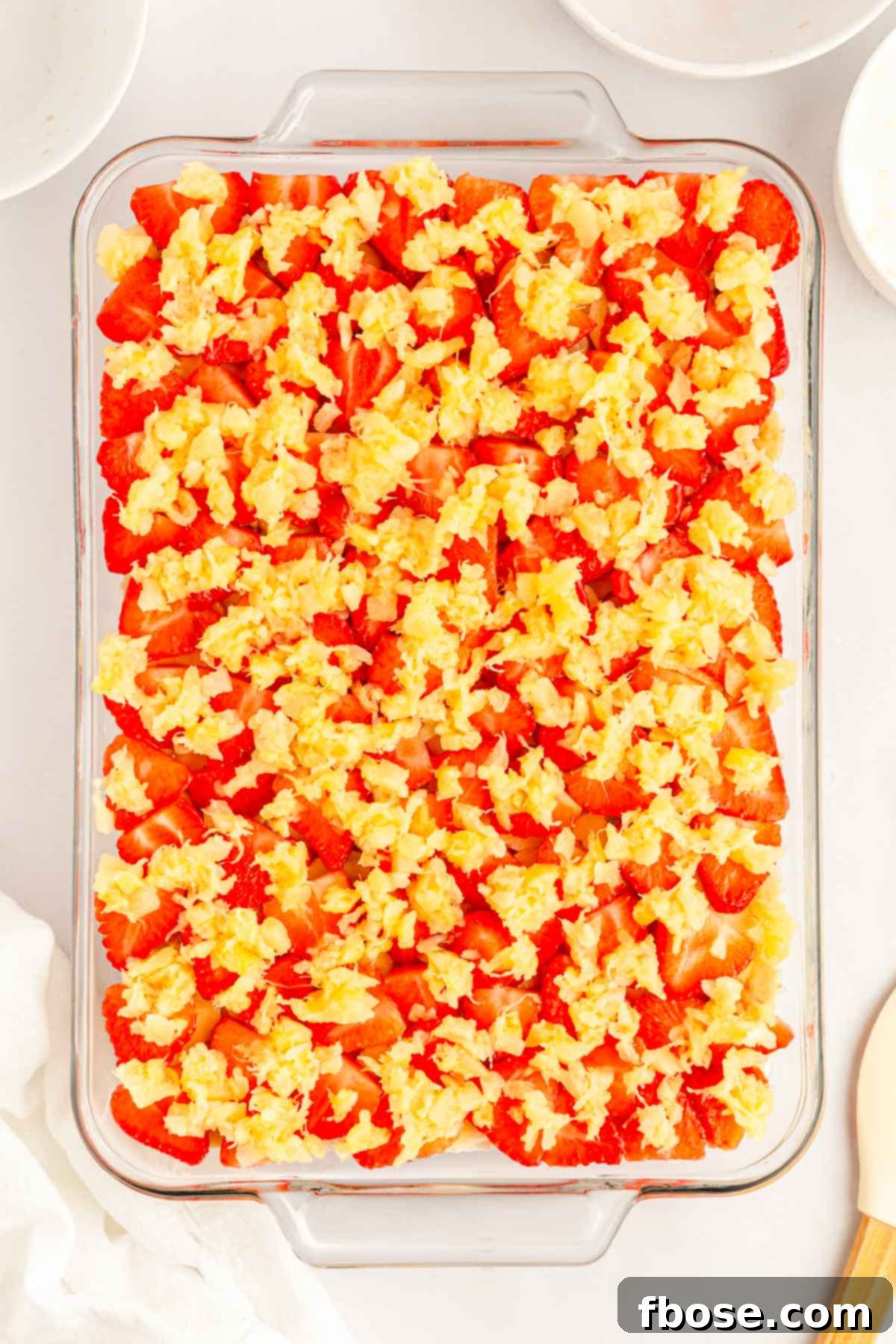 Colorful layers of sliced bananas, strawberries, and crushed pineapple arranged in a glass baking dish, sitting atop the creamy filling.