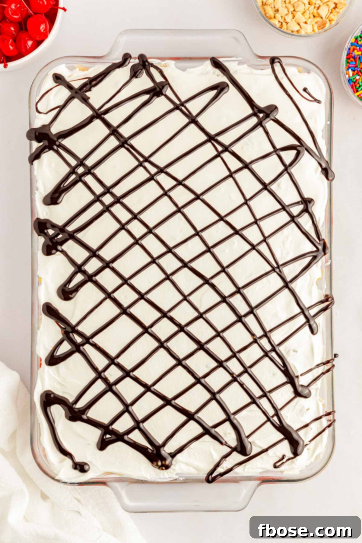 A generous layer of whipped cream (Cool Whip) spread over the fruit, with a drizzle of chocolate syrup on top, in a glass pan.
